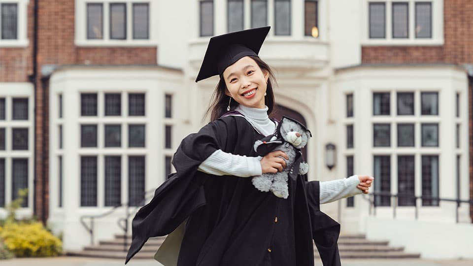 Xu wearing a graduation gown and cap holding her degree scroll outside Hazlerigg Building on Loughborough University campus. She is smiling and she is also holding a grey stuffed teddy bear which is also wearing a black cap and gown.